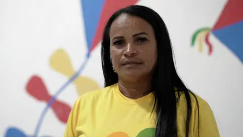 Felix Lima/BBC News Brasil Alessandra Hora looks at the camera. She is wearing a yellow t-shirt and has long black hair.