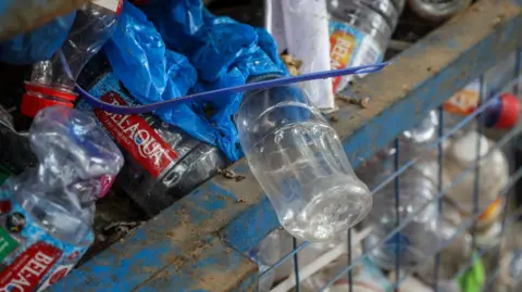 Nipah Dennis/Bloomberg/Getty Images A blue metal gridded bin is stuffed full of plastic bottles of different colours. At the top of the bin a clear plastic bottle hangs over the side of the bin, on top of it is blue plastic and a blue cable tie