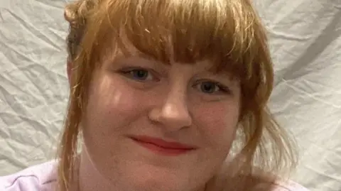 Maisy Moazzenkivi Maisy Moazzenkivi with her short red hair tied up smiling at the camera and wearing a pink t-shirt