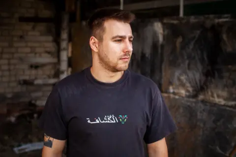 Stanislav Ostrous/BBC Dmytro Olifirenko wears a dark T-shirt with a scrawled logo in white and a tattoo partially showing on one arm