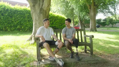 TOBY RONEY Two individuals, one with a prosthetic leg, sitting on a wooden bench in a park, surrounded by trees.

