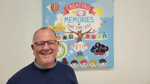 The Entertainer Gary Grant pictured today wearing glasses, a checked shirt under a dark blue jumper, standing in front of a poster that says "Creating memories that can last a lifetime" illustrated with a tree and drawings of children with thought bubbles showing pictures of a castle and outer space