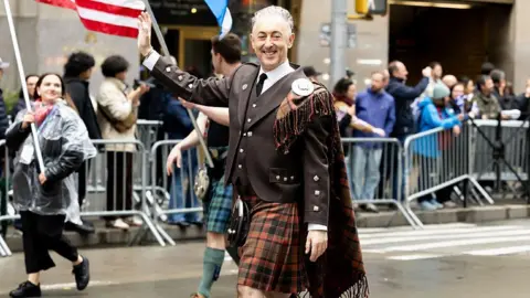 Getty Images Alan Cumming, in brown and soft red Highland suit and kilt, is smiling and waving while walking along the middle of the street in New York. People are cheering in the background.