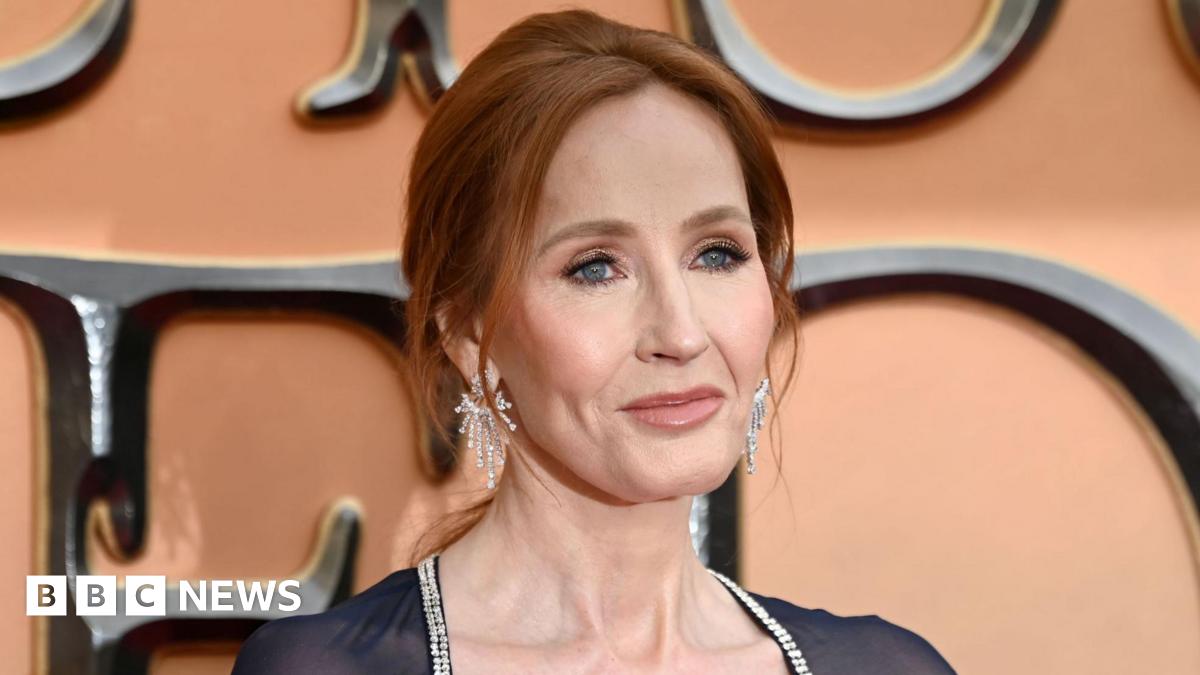 Head shot of Harry Potter author JK Rowling, She has red hair, tied back in a ponytail and is wearing long diamond earrings. She is wearing a black dress, with a diamond trim. Rowling is standing in front of a pink backdrop advertising "Fantastic Beasts: The Secret of Dumbledore". She is looking off to the left of the camera.