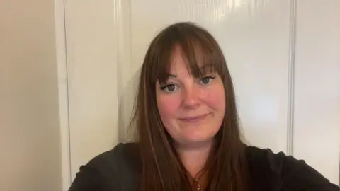 Jemma Caffrey Jemma stands in a hallway looking into the camera. She has brown hair and a black top.