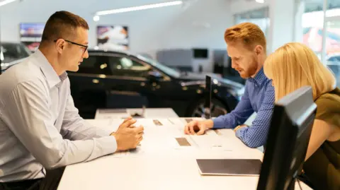 Getty Images Salesperson wearing a white shirt advising couple in car dealership as they sit around a table, with a black car in the background