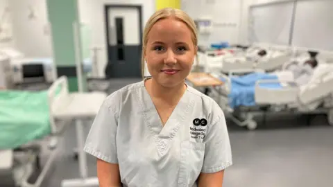 BBC/ Kate McGough Freya has blonde hair tied back and wears a uniform with her college branding. Behind her, out of focus, is a mock-up hospital room in a college, with mannequins lying in hospital beds.