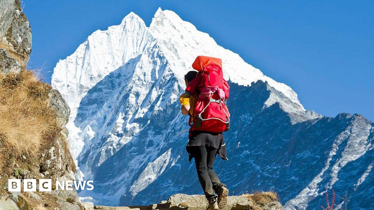 A woman carrying her toddler child in a red child carrier rucksack on her back, trekking above Namche Bazar (3,440 m), the base for trekking and mountaineering in Nepal's Solo Khumbu region