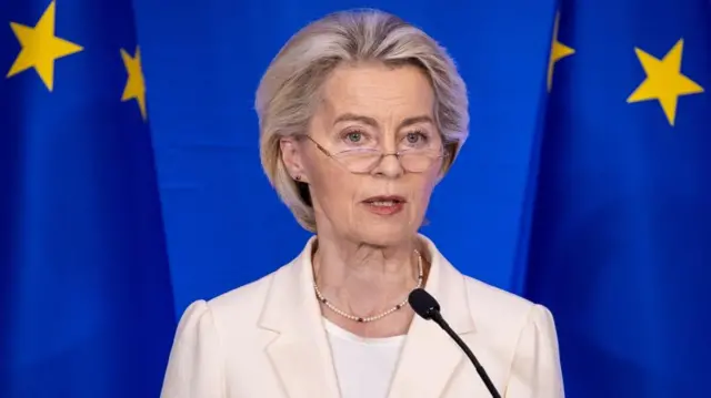 Ursula von der Leyen in a white blazer and t-shirt with a beaded necklace. She stands behind a microphone while speaking, she is standing in front of a large blue curtain with yellow stars on it.
