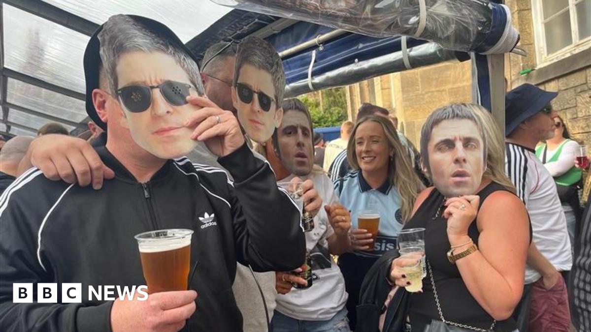 A group of Oasis fans. A man on the near left is wearing a black adidas track top with a white logo on the upper right chest and three white stripes on the arms. He is holding a face mask of Noel Gallagher to his face in his left hand. A man next to him has the same mask attached to his face. On the far side, a woman holding a cup of beer is wearing an oasis T-shirt. A woman next to her wearing a black top is holding a cup of wine in one hand and a face mask of Liam Gallagher to her face with the other.