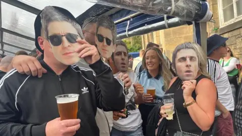 BBC A group of Oasis fans. A man on the near left is wearing a black adidas track top with a white logo on the upper right chest and three white stripes on the arms. He is holding a face mask of Noel Gallagher to his face in his left hand. A man next to him has the same mask attached to his face. On the far side, a woman holding a cup of beer is wearing an oasis T-shirt. A woman next to her wearing a black top is holding a cup of wine in one hand and a face mask of Liam Gallagher to her face with the other.