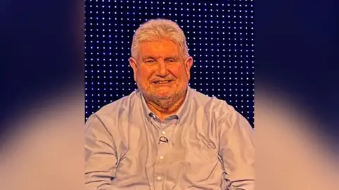 ITV Tim McCarthy has thick white hair and is wearing a light grey open-necked shirt. He is smiling with coloured lights seen behind. 
