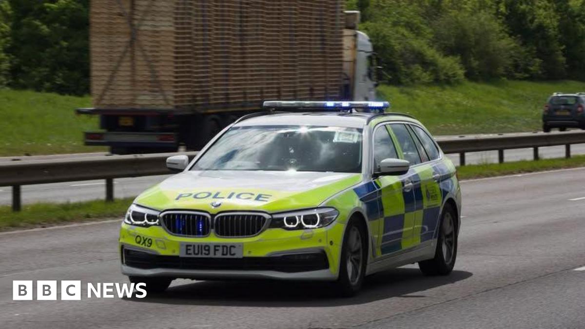 A police car on a motorway with a lorry carrying pallets in the background driving in the opposite direction.