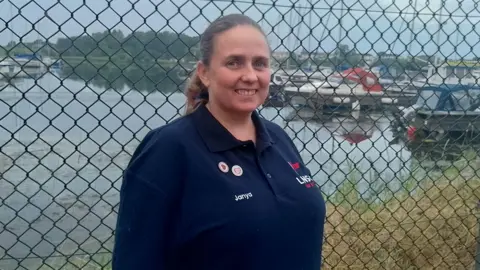 A woman with long brown hair, tied back. She is wearing a navy t-shirt. She's standing in front of a fence, with Lough Neagh visible in the background. 