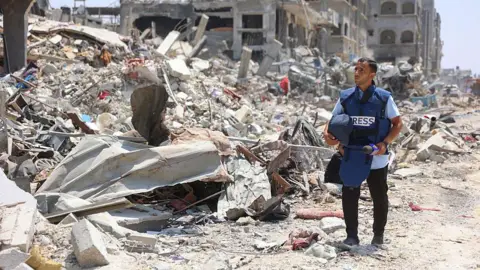 AFP via Getty Images Anas al-Sharif stands wearing a "press" vest and holding a helmet in one hand as he surveys the destruction done to buildings around him in Gaza City. To his left is a huge pile of rubble and debris. 