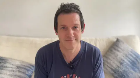 A man with short brown hair and a blue t-shirt sits on a cream sofa, looking directly into the camera. Behind him is a white wall, on the sofa there are blue cushions, one either side of him. 
