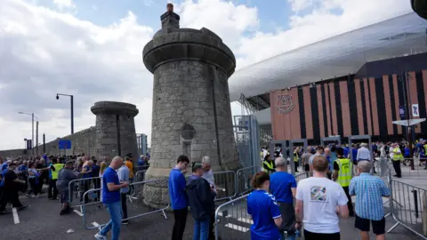 PA Media Crowds of fans, mainly in blue clothing, line up in queues leading to security gates that are next to tall dock towers made of grey granite stone. The old dock wall continues around the stadium behind. The arena has a striped orange and black front with the Everton logo under a silver metal roof.
