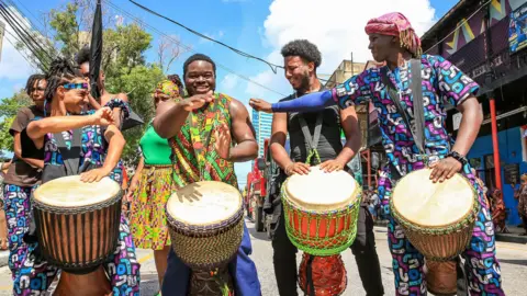 EPA/Shutterstock Four youngsters play the drums. They are stood in a row and dressed in bright, patterned clothing.