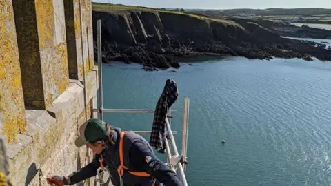 Mike Conner Scaffolding set up along one of the fortress walls, with a man on top of it using a hand tool. He is wearing a cap, blue gilet and orange harness. Behind the scaffolding is the sea, with cliffs in the background. 