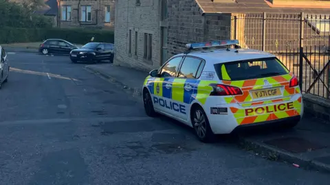 BBC A police car is parked on the pavement alongside an iron fence.