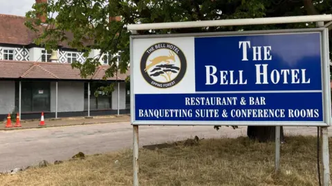The Bell Hotel, a white building, is on the left with an entrance to the right which says The Bell Hotel, Best Western. A tree is in front of the two-storey building with three traffic cones outside.