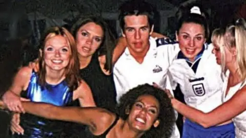 Anthony Kavanagh Group image of Geri Halliwell in a blue dress and Victoria Adams in a black dress on the left of Anthony in a white shirt. mel C wears a white England football top with Emma Bunton in white at the right. Mel B crowches down in front stretching out her arms.