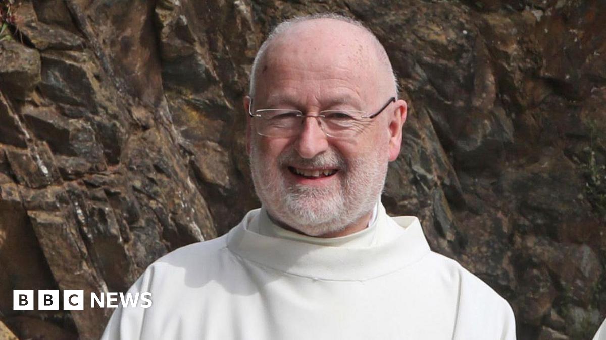 Fr John Murray. He is smiling at the camera. Fr Murray is bald and is wearing glasses. He has a white beard and is wearing white robes. Behind him is a rockface.