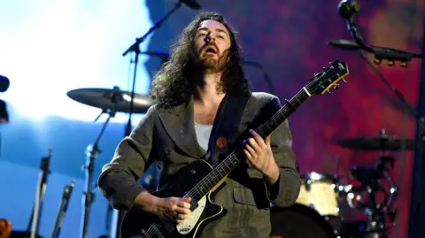 Getty Images Hozier who has long curly hair and green jacket playing an electric guitar on stage. There is a set of drums behind him with a bright blue and dark red background.