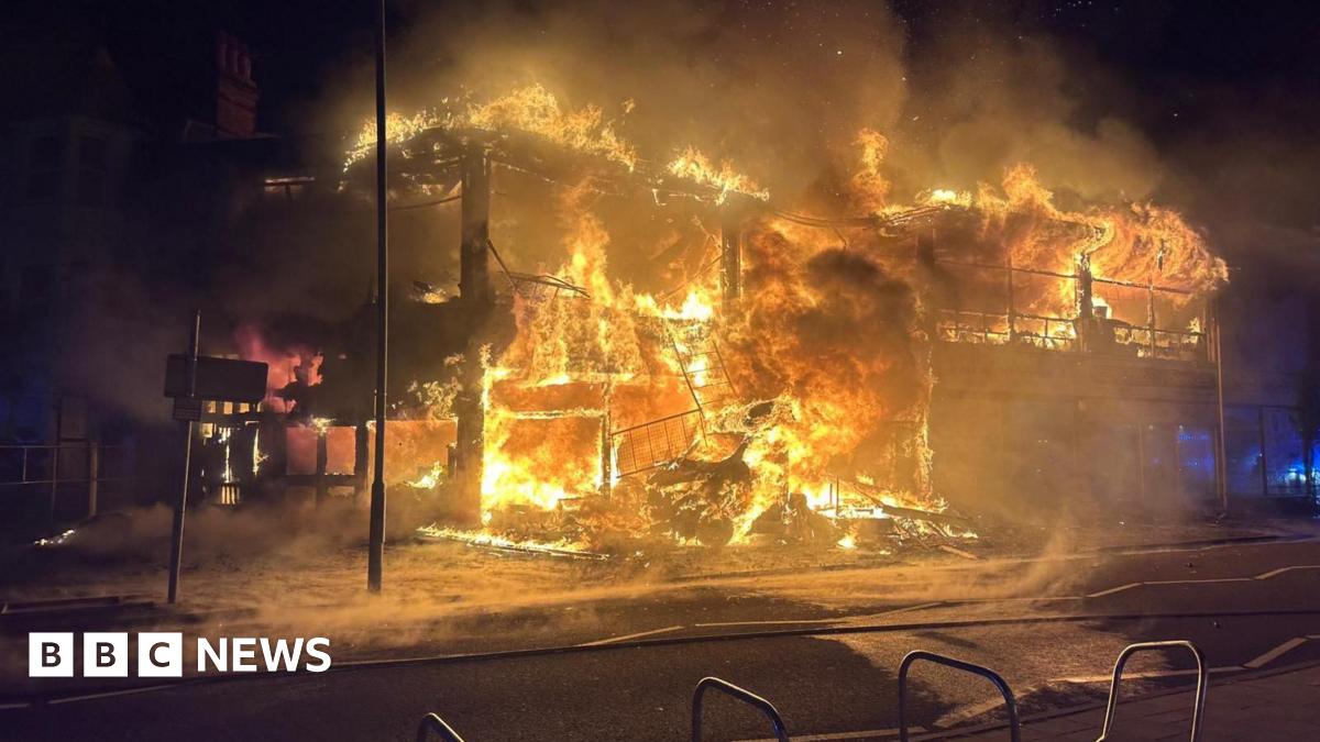 A fire is earing through a two storey building and has burnt down the walls, just leaving the bones and foundations of the site. It remains engulfed in flames. The sky is dark making the fire appear even brighter against the night sky.
