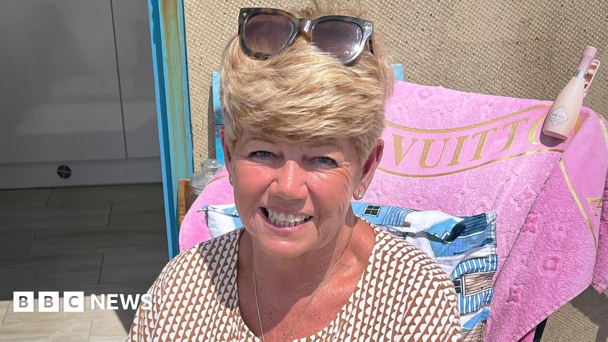 Debbie Booker is sitting outdoors. She has short blonde hair and is wearing sunglasses on top of her head and a patterned shirt. Behind her, a pink towel with the word "VUITTON" is draped over a chair. A bottle is placed on the chair's armrest. The background shows part of an open door and tiled flooring.