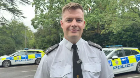 A police officer in a white shirt with a black tie and black shoulder pads. He is stood in front of two police cars.