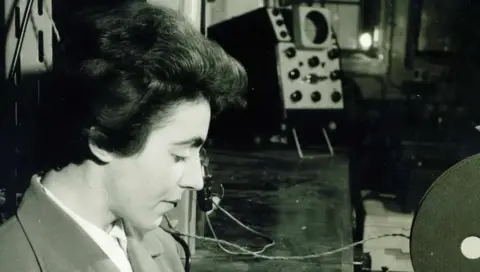 Dame Stephanie Shirley Dame Stephanie is pictured in a black and white photograph looking down and wearing a shirt and blazer with technological equipment in the background