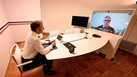 Getty Images Emmanuel Macron sits at a table in front of a video conferencing screen. Macron has short brown hair and wears a white shirt and dark suit trousers. He is sat at a round white table in a white-walled room with brown tiled flooring. At the other end of the table is a large screen on which a video conference is being held. Sir Keir Starmer is enlarged on the screen, while a row of small boxes containing the other participants sits below him. Starmer wears a grey top and black-rimmed glasses, and sits in front of a plain white background.