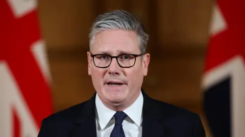Reuters Keir Starmer. He has short grey-ish hair, wearing glasses, white shirt and dark blazer and tie. Two British flags are in the background.