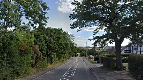 Google Cowley Hill in Borehamwood showing a single carriageway road going down a hill towards a mini roundabout. Trees are to the left and some homes on the right, with a big tree on the right pavement.