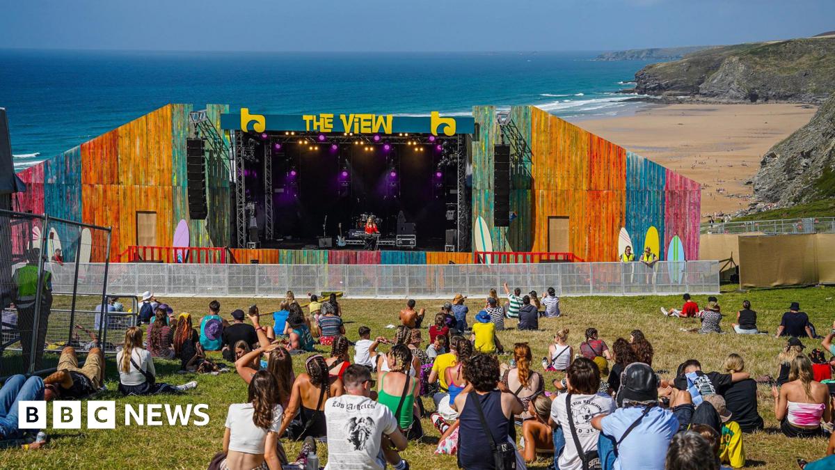 A colourful outdoor stage labelled 'THE VIEW' set up on a grassy area overlooking a beach and the ocean. The stage features vibrant multi-coloured panels and purple lighting. A small crowd sits on the grass watching a performance, with cliffs and the shoreline visible in the background.