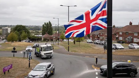 Getty Images A Union Jack flag hangs from a lamppost above a roundabout, as cars pass beneath it and houses and other flags hanging from lampposts can be seen in the distance.