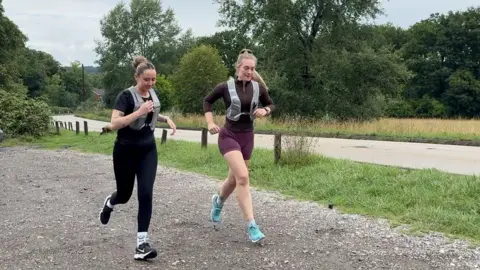 BBC Image shows two females running. One is dressed in all black. She has a black short-sleeved top and black leggings on with a grey running vest over the top. She has black Nike trainers on. Her hair is in a bun. The second officer has turquoise trainers on. Burgundy shorts and a brown long-sleeved tight fitting top. She has a grey running vest over the top. Her hair is in a ponytail. They are in a carpark with four cars parked alongside them. Behind them are a number of trees, grass and a main road. 