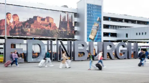 Getty Images Exterior image of Edinburgh Airport, featuring a large Hollywood-style sign with "EDINBURGH" in grey block capital letters. A number of blurred passengers can be seen walking across the concourse outside the terminal building.