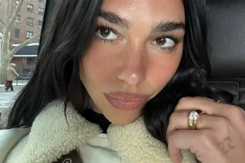 @dualipa/Instagram Selfie image of Dua Lipa, where her left hand is on show with her engagement ring - a thick gold band with a diamond in the centre - on show.