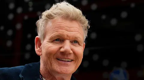 Getty Images Gordon Ramsay, a man with short blonde hair wearing a black zip-up jacket over a white t-shirt. He is smiling in front of a blurred black background.