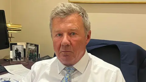 Eric Masterson sits at a desk in an office, wearing a white shirt and a blue floral-patterned tie.  A dark blue jacket is draped over the chair behind him. The desk holds papers, documents, and two framed family photographs. A computer monitor and a brass desk lamp are visible in the background.