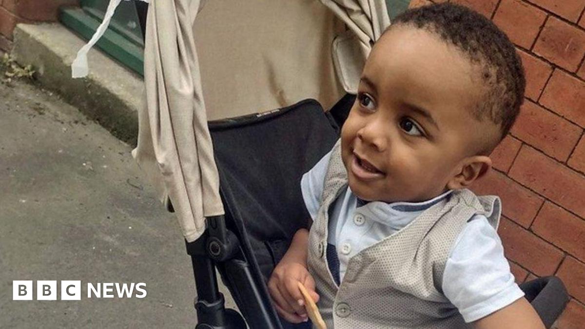 Family photo of Exodus Eyob, a small boy looking to the side of the camera while sitting in a black pushchair with a cream hood. He has short dark hair and is wearing a white t-shirt with buttons at the neck and a cream waistcoat, and is holding a biscuit. He is pictured outside with a brick wall behind him.