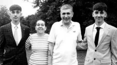 Thetford Town Football Club/Facebook The family of four are standing outside with their arms around each other. They are all facing the camera. There are three men, two siblings and and older man, who are a similar height. Between them is a women who is much smaller. They are all smiling and the picture has been taken in black and white.