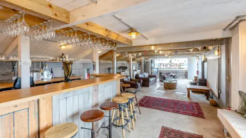 Strutt & Parker A bar area created in the fortress. The bar itself is wooden, with wooden and metal bar stools and wine glasses hanging from the ceiling on wire racks. Beyond, Afghan rugs cover parts of the concrete floor, with leather armchairs and a window looking out to the sea. 