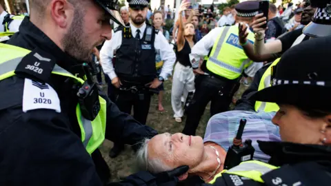 EPA A woman with grey hair and wearing a necklace and checked shirt is carried away by officers, one of whom is holding her head as she lies still. In the background more officers and people taking pictures can be seen