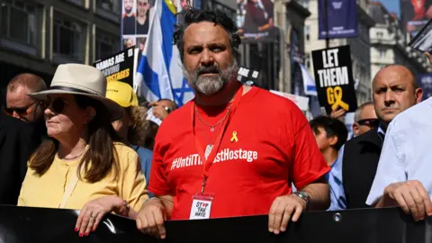 Reuters Adam Ma'anit, a man with short black hair and greying facial hair wearing a red t-shirt, walks at the front of a large protest with an Israeli flag and several placards in the background, including one which says 'let them go'.