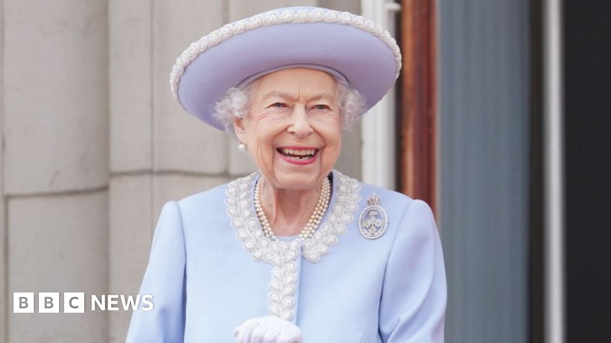 The late Queen Elizabeth II smiles broadly as she stands outside, wearing a lilac hat and jacket - her white gloves can just be seen and she has several strands of pearls around her neck