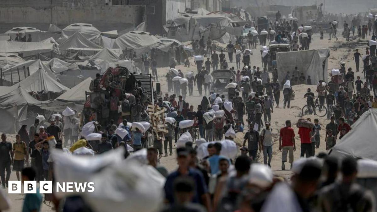 A landscape image of a dirt track with a number of people walking and carrying white aid packages. Tents can also be seen.