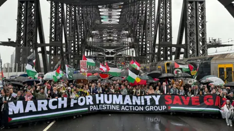 Dean Lewins/EPA Hundreds of protesters gather under the arches of the Sydney Harbour Bridge, wearing raincoats and holding red, green, black and white flags. They are holding a large sign that reads "March for Humanity: Save Gaza".